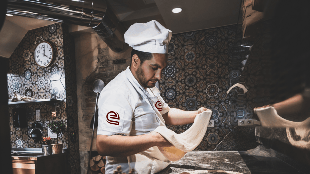 A person in branded chef's uniform forming dough in a kitchen.