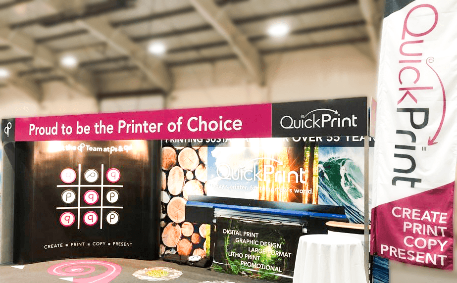 A photo of QuickPrint's trade show display stand, complete with header and display boards, a vertical flag, and even a poster printer